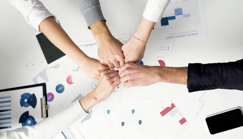Team von Business-Intelligence-Experten stößt Fäuste zusammen über Schreibtisch mit Diagrammen, KPIs und Analyseberichten – Symbol für datenbasierte Entscheidungen, transparente Geschäftsprozesse und skalierbare BI-Architektur im DACH-Raum.